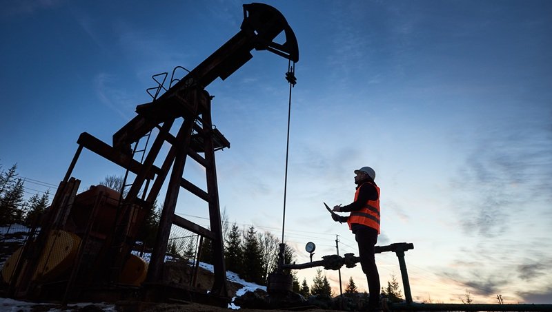 Oilfield operations at dusk Worker standing beside an operating oil pump jack at dusk
