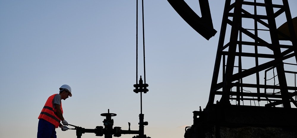 Oilfield worker in a red hard hat servicing a pump jack wellhead at a U.S. oil and gas site